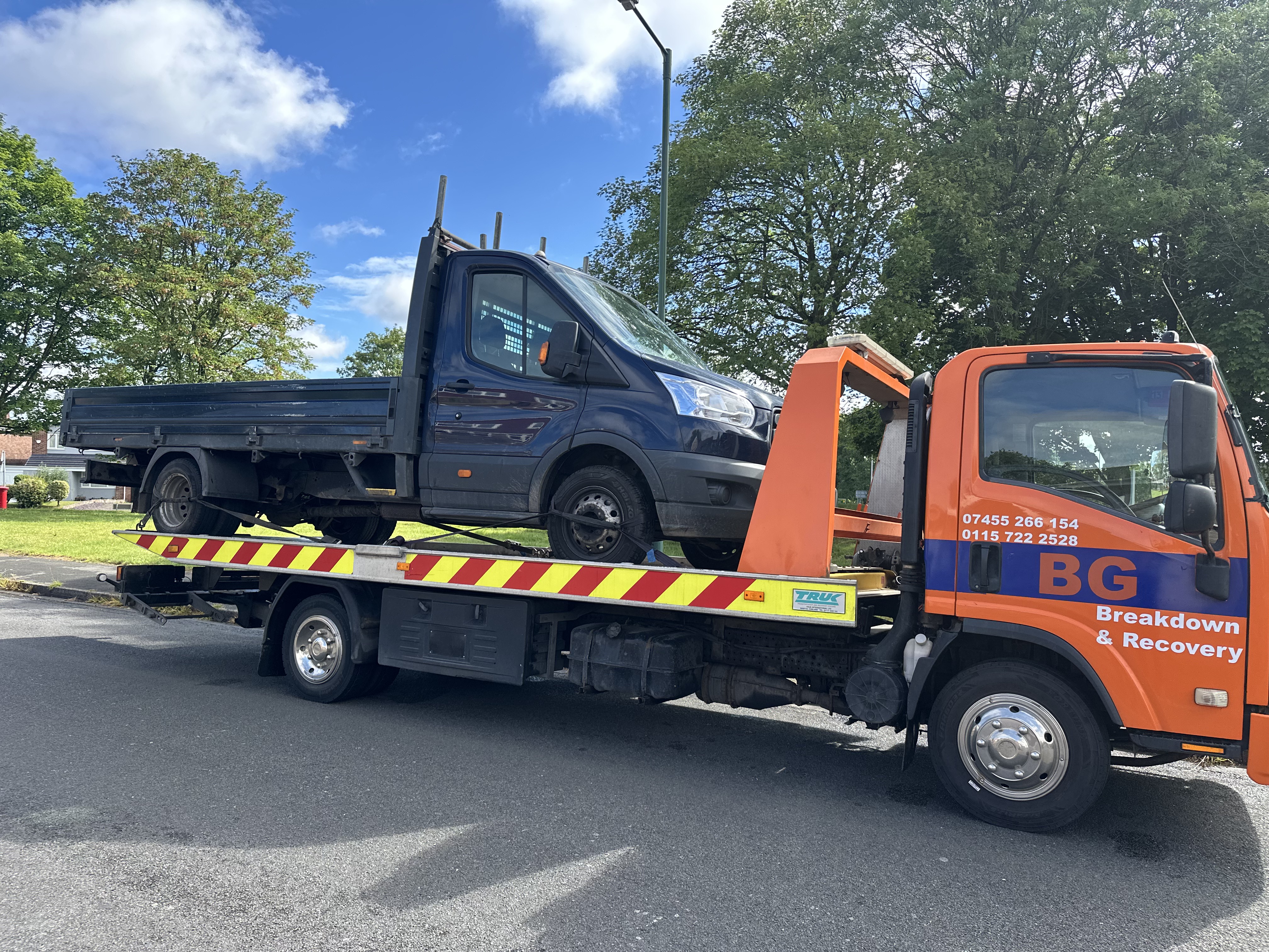 A flatbed tow truck transports a small utility pickup truck or van on the back of the truck. The truck is orange with a bl...